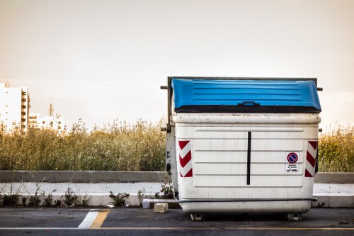 A large commercial skip with a white body and a blue lid is positioned on a paved surface, likely a parking area or outdoor space. The skip has red and white diagonal reflective markings on its sides and a small round no-parking sign adhered to the front. It is situated close to a curb, with some low-growing grass and weeds growing along the edge of the pavement. In the background, there is a stretch of natural greenery, including tall grass or bushes, under a muted, overcast sky. The scene is minimally populated, focusing on the skip as the primary object, with an environment that suggests it is used for rubbish or garden clearance in an urban or suburban setting, possibly within the Shadwell area, consistent with the services offered by Garden Clearance Shadwell. The overall atmosphere is quiet and utilitarian, fitting for rubbish removal contexts in the local area.