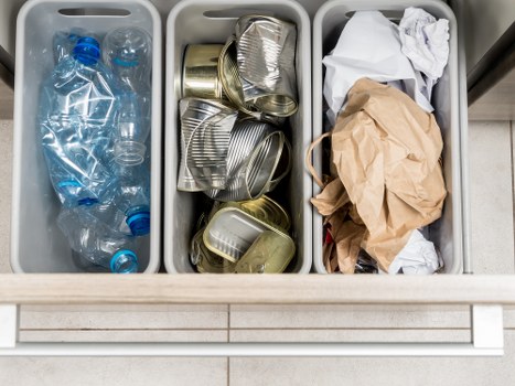 A three-compartment recycling bin positioned on a tiled floor, with the left compartment containing clear plastic bottles, some with blue caps, partially crushed. The middle section holds several used aluminium food and drink cans, some flattened and stacked, with visible ridges and metallic sheen. The right compartment contains crumpled brown paper and white tissue paper, with the paper appearing loosely bunched and the tissues loosely folded. The bin is part of an outdoor or semi-outdoor setting, suggesting it is used for disposing of household waste and recyclables as part of rubbish removal services, possibly in a residential area near Shadwell or the London postcode area. The organized sections aim to separate plastics, metals, and paper waste for appropriate disposal or recycling, and the image illustrates the typical contents handled by Garden Clearance Shadwell for professional rubbish removal and garden clearance tasks.